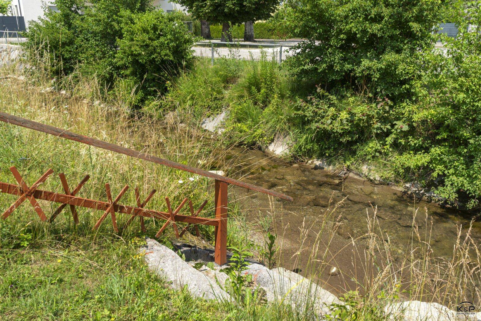 Kleiner Bachlauf mit Uferbewuchs und rustikalem Geländer neben dem Grundstück.