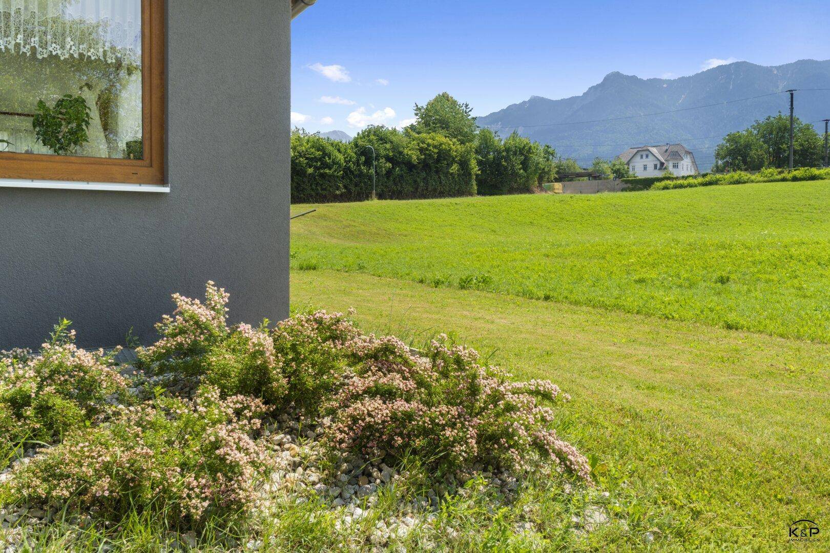 Blühender Strauch neben dem Haus mit weitem Blick auf grüne Felder und Berge.