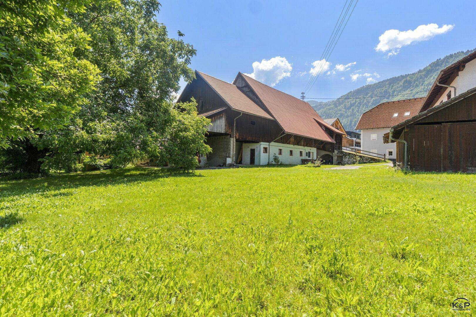 Große grüne Wiese mit traditionellem Bauernhaus im Hintergrund und sonnigem Bergblick.