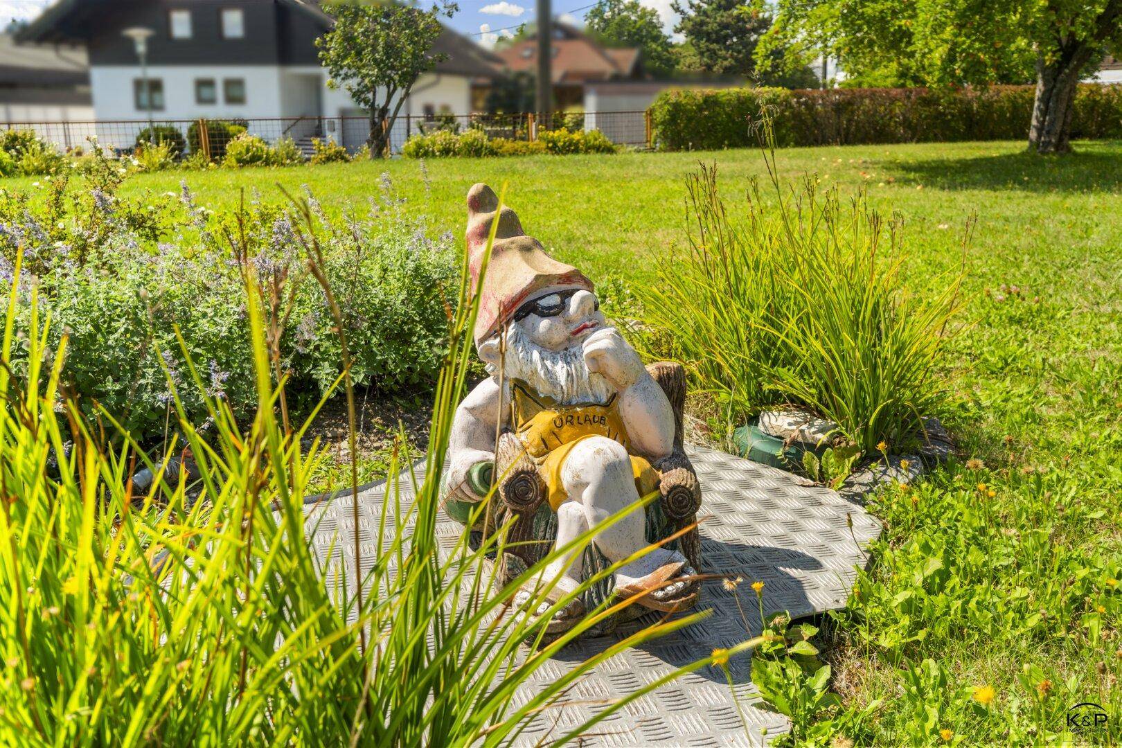 Ein Gartenzwerg sitzt nachdenklich auf einem Stuhl in einem sonnigen Gartenbereich mit viel Grün.