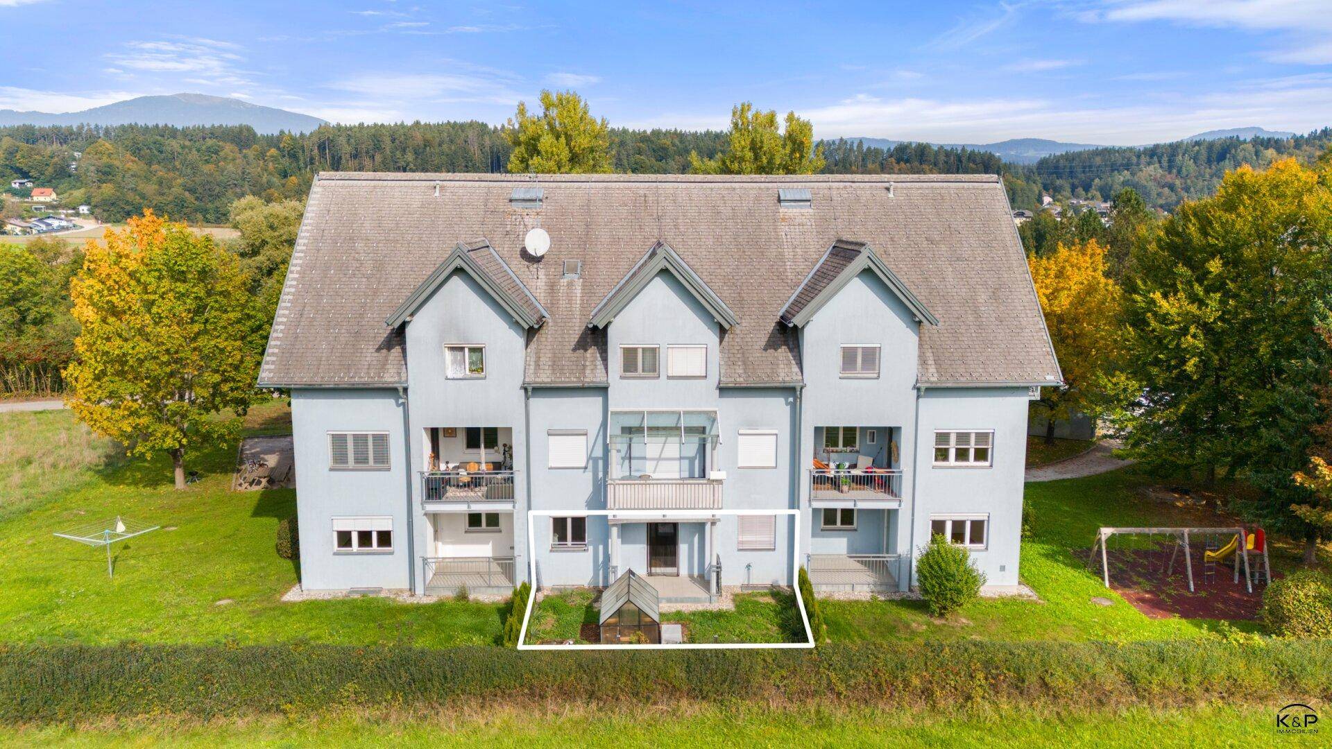 Ansicht des hellblauen Mehrfamilienhauses mit Balkonen und einem kleinen Spielplatz im Grünen.