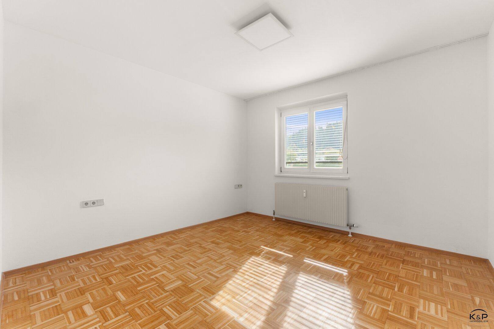 Gemütliches Schlafzimmer mit Parkettboden, weiß gestrichenen Wänden und einem Fenster mit Jalousien.