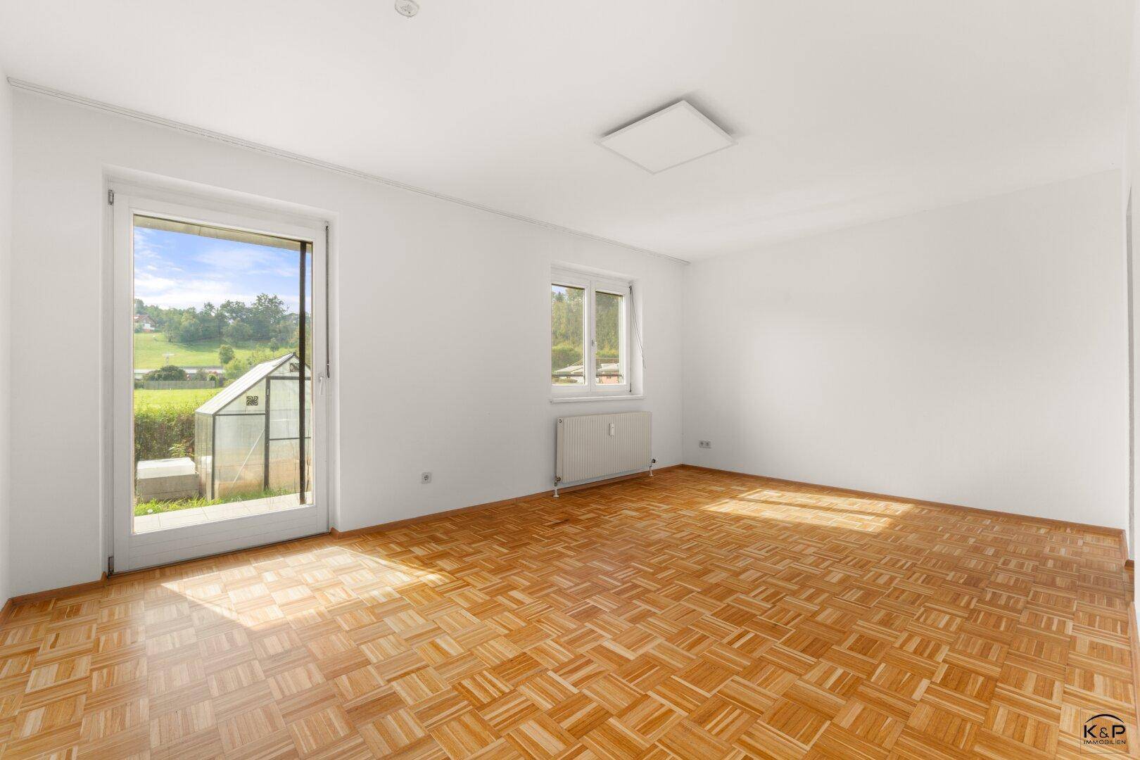 Heller Wohnraum mit Parkettboden, großen Fenstern und direktem Zugang zum Garten mit Gewächshaus.