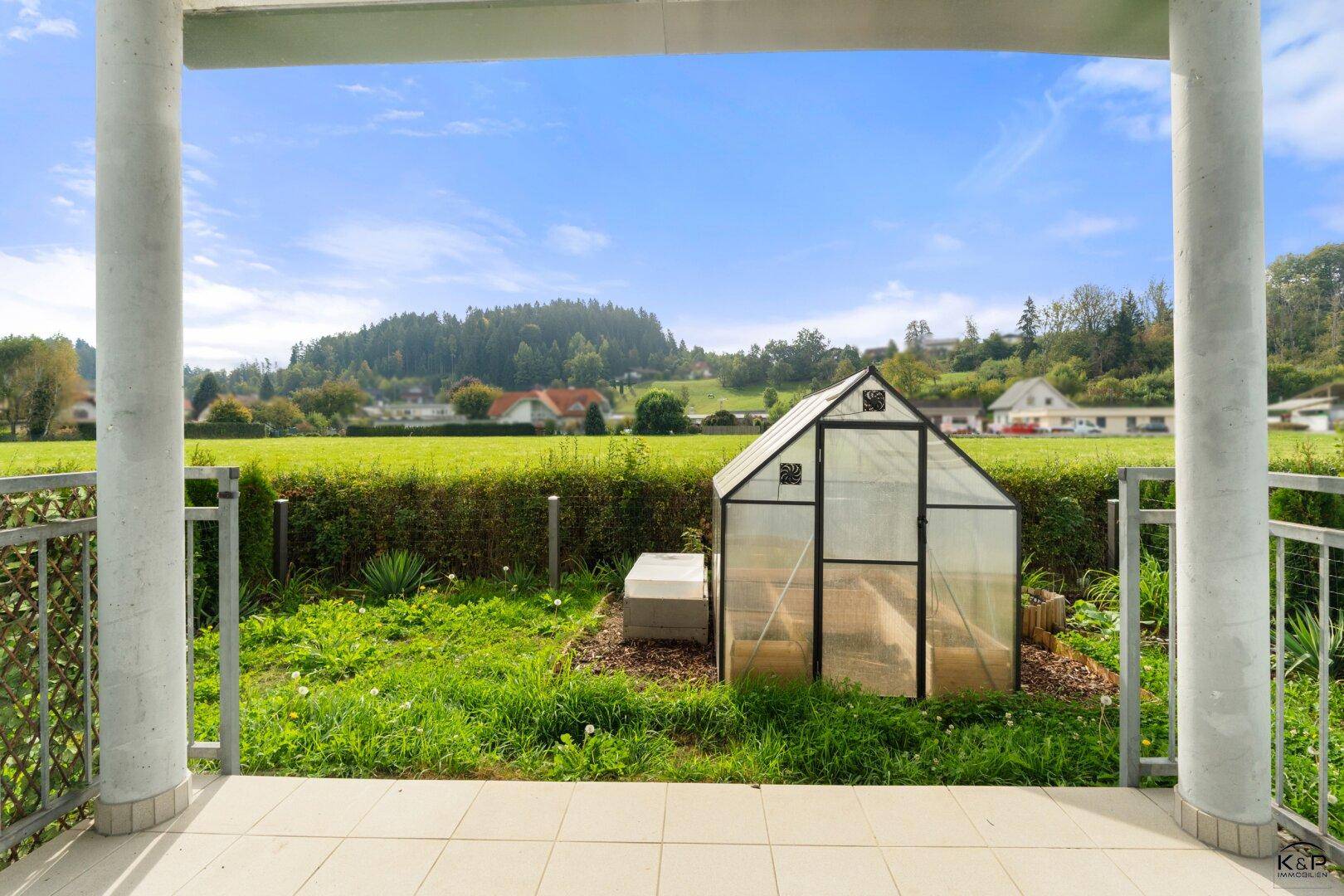 Überdachte Terrasse mit Blick auf den kleinen Garten, ein Gewächshaus und weite grüne Felder.