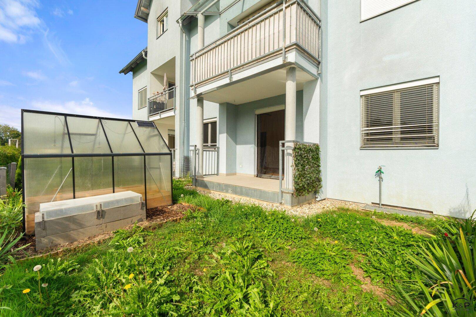 Erdgeschosszugang mit kleinem Gartenbereich, Gewächshaus und der hellblauen Gebäudefassade.