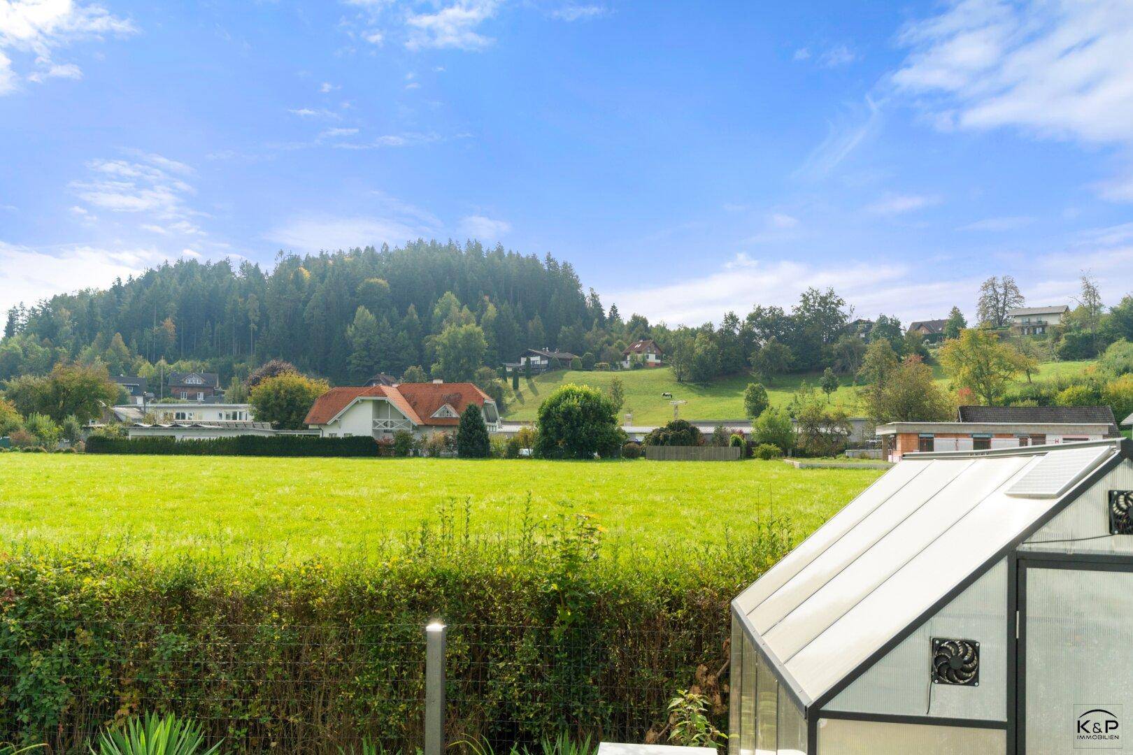 Weitblick vom Garten über das Gewächshaus auf grüne Felder und die umliegende ländliche Bebauung.