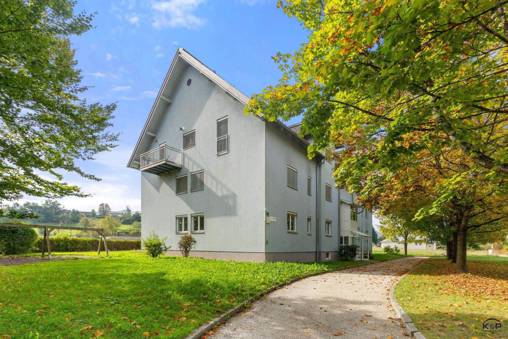 Seitenansicht des Mehrfamilienhauses mit hellblauer Fassade, umgeben von herbstlichen Bäumen und Grünflächen.