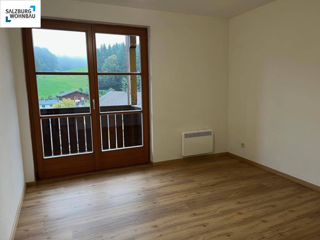 Helles Zimmer mit Laminatboden und Zugang zu einem kleinen Balkon mit Blick ins Grüne.