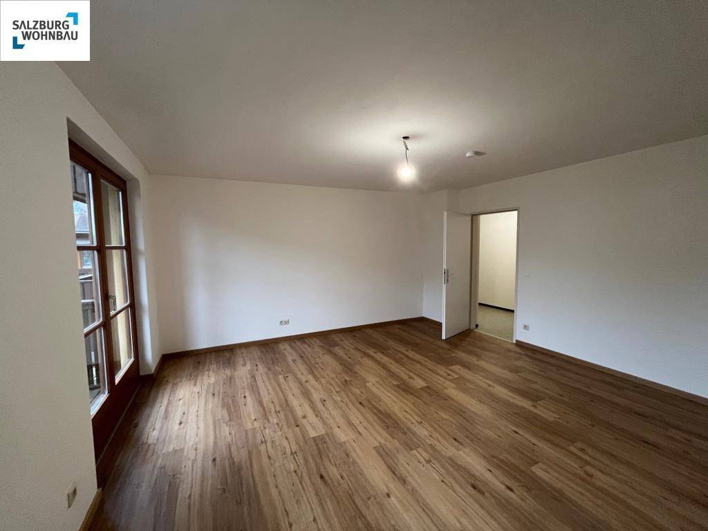 Geräumiges Wohnzimmer mit Laminatboden und hellem Fenster, das viel Tageslicht hereinlässt.