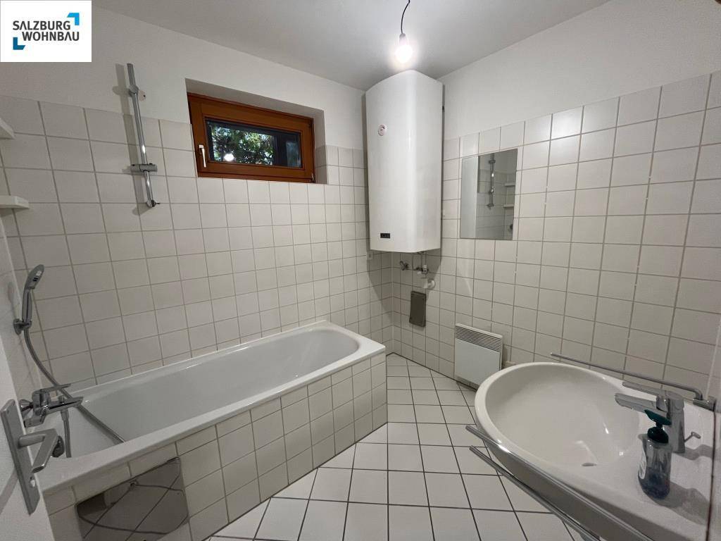 Badezimmer mit Badewanne, Waschbecken und weißen Fliesen, inklusive Fenster für Tageslicht.