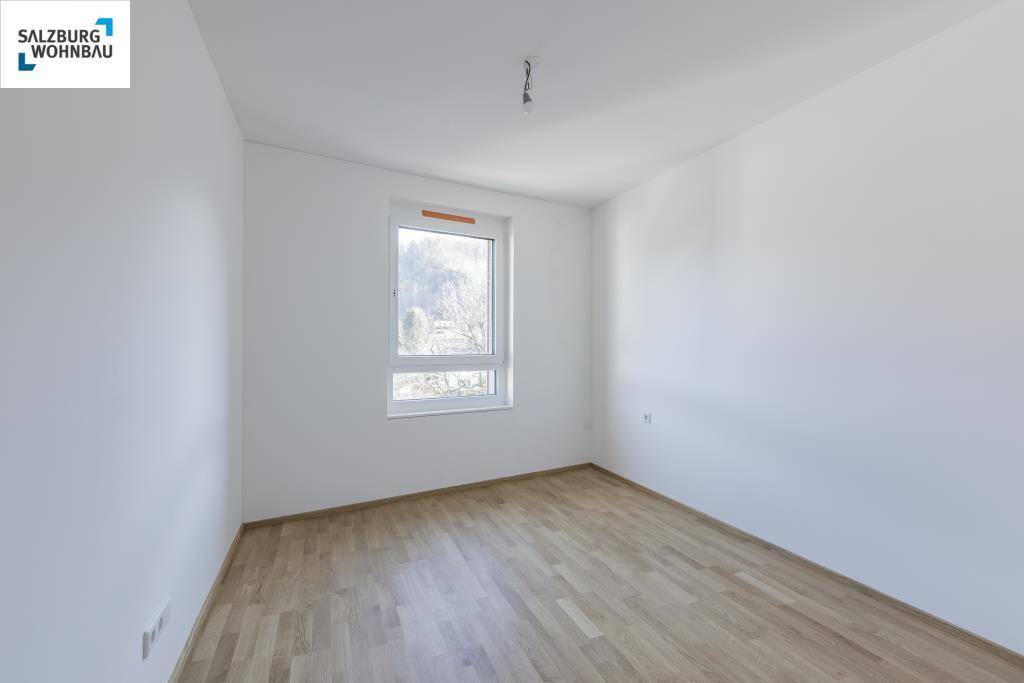 Helles, unmöbliertes Schlafzimmer mit Holzboden und einem Fenster mit Blick ins Grüne.