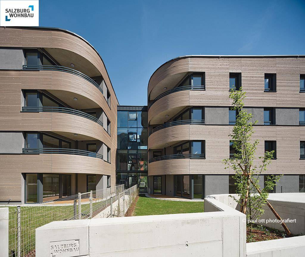 Zwei moderne Wohngebäude mit geschwungenen Holzfassaden und Balkonen, umgeben von Grünflächen.