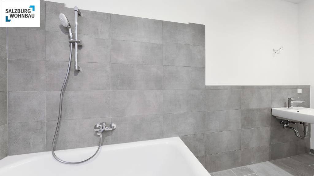 Helles Badezimmer mit grauen Fliesen, Badewanne mit Duschfunktion und einem einzelnen Waschbecken.