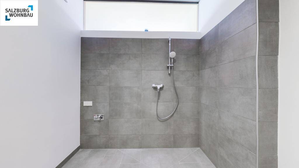 Modernes Badezimmer mit grauen Fliesen, Duschbereich und einem hohen Fenster für Tageslicht.