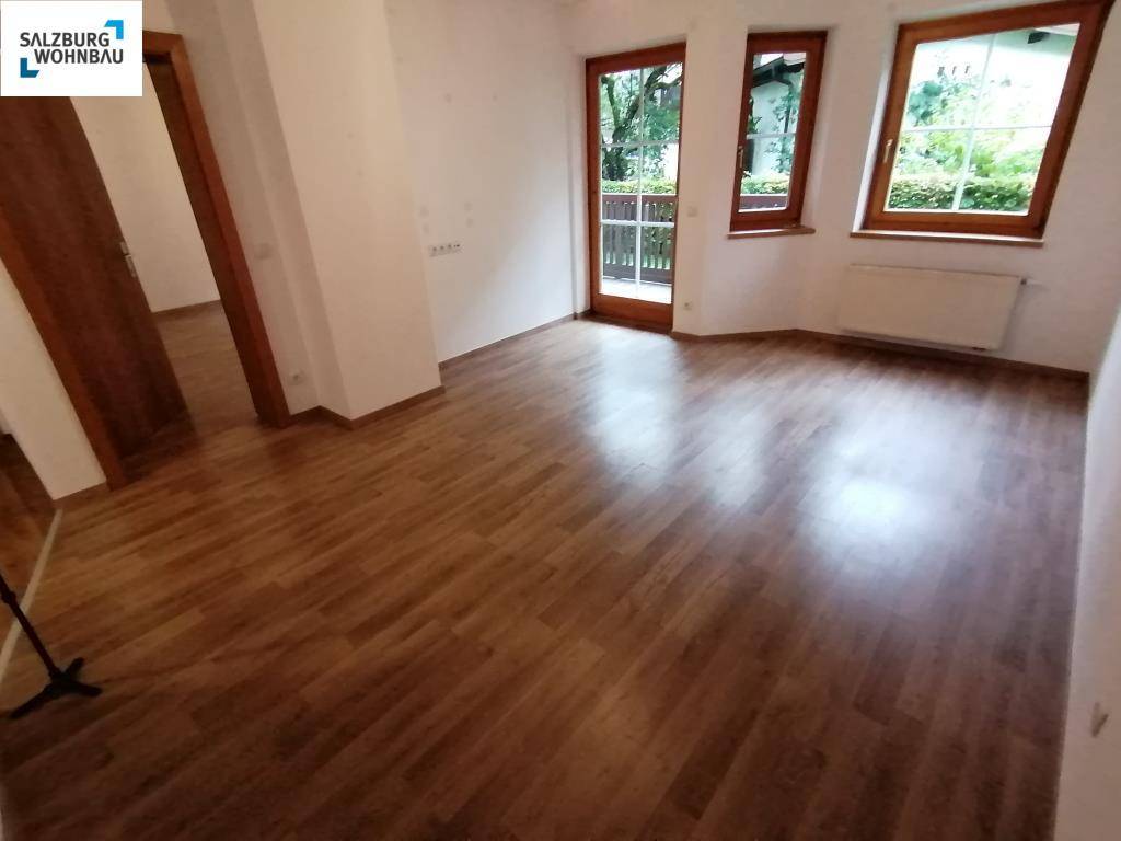 Heller, leerer Wohnraum mit Laminatboden, großen Fenstern und Zugang zum Balkon.