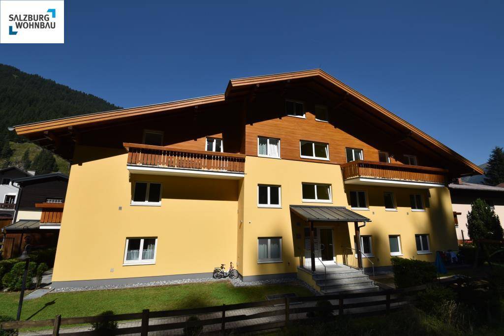 Gelbes Wohngebäude mit Holzbalkonen und traditionellem Satteldach vor bewaldetem Berghang.