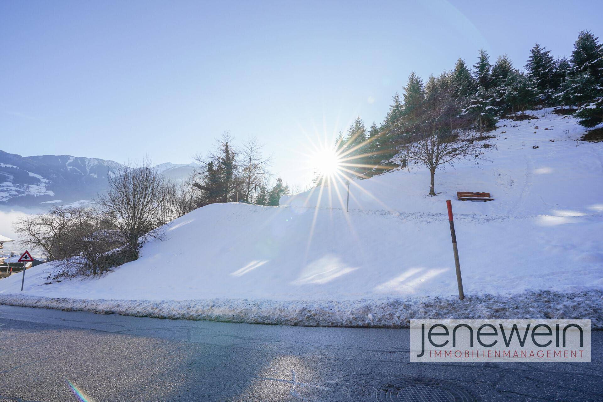 Sonnige Winterlandschaft mit verschneitem Hang, Straße und Blick auf die Berge im Hintergrund.