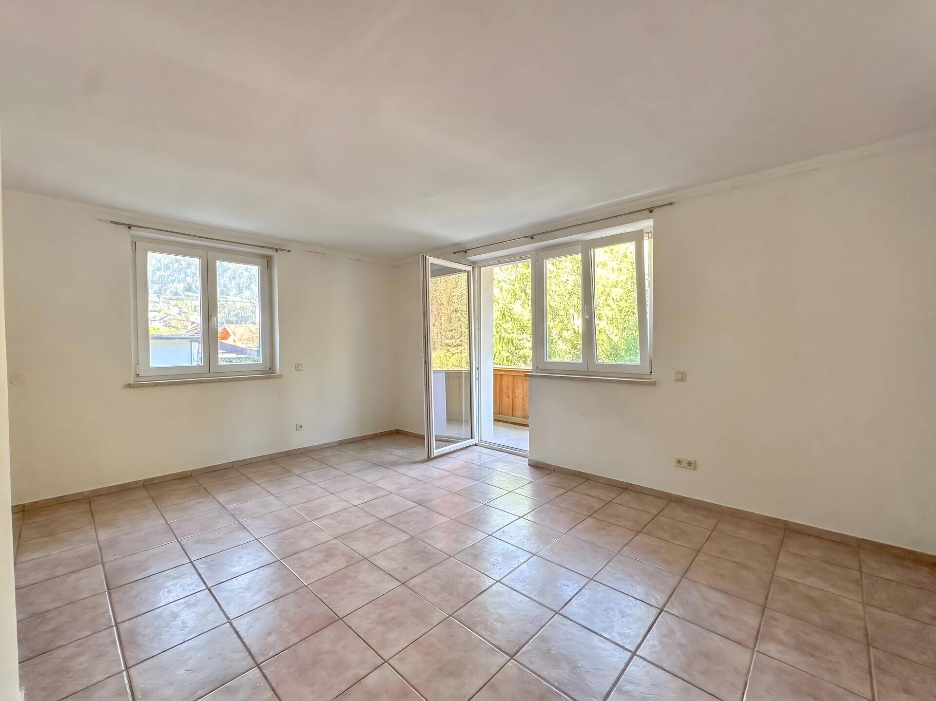 Helles, leeres Wohnzimmer mit Fliesenboden, großen Fenstern und Zugang zum Balkon.