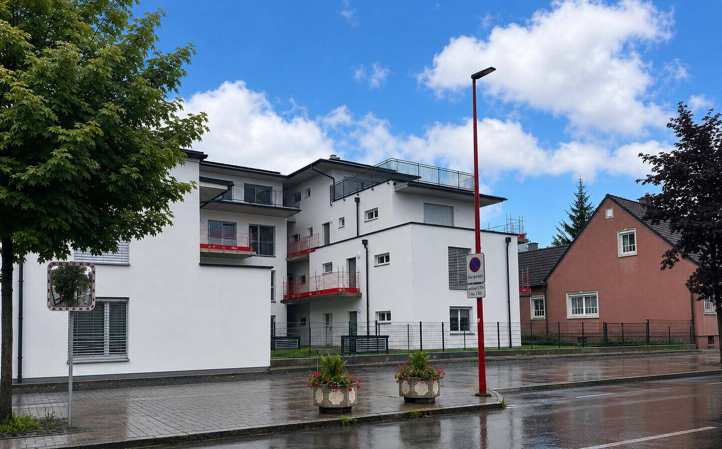 Seitenansicht des modernen Wohnhauses mit roten Balkonen, weißer Fassade und umliegender Bebauung.
