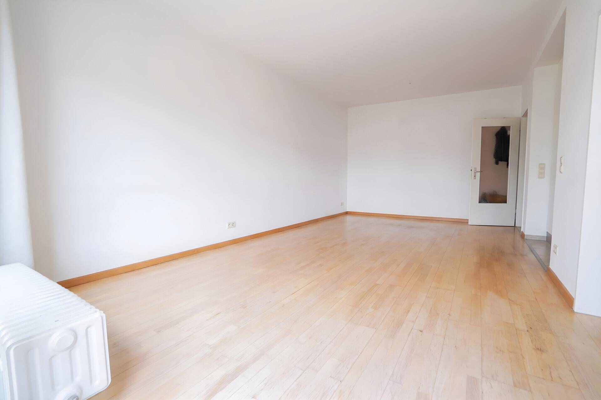 Helles, leeres Wohnzimmer mit hellem Holzfußboden und weißen Wänden.