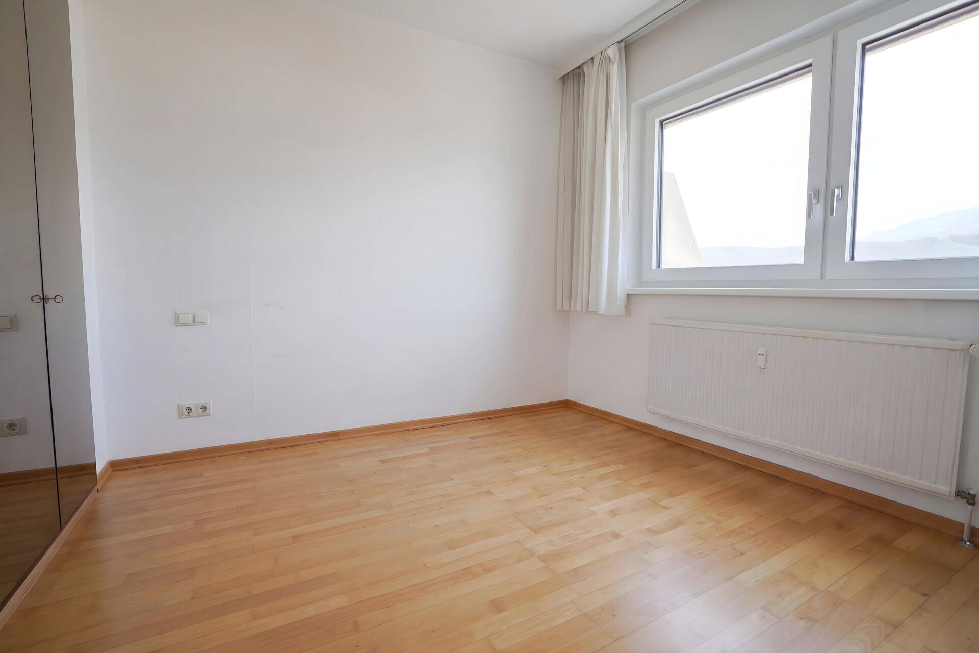 Leeres Schlafzimmer mit hellem Holzfußboden, weißen Wänden und großem Fenster.