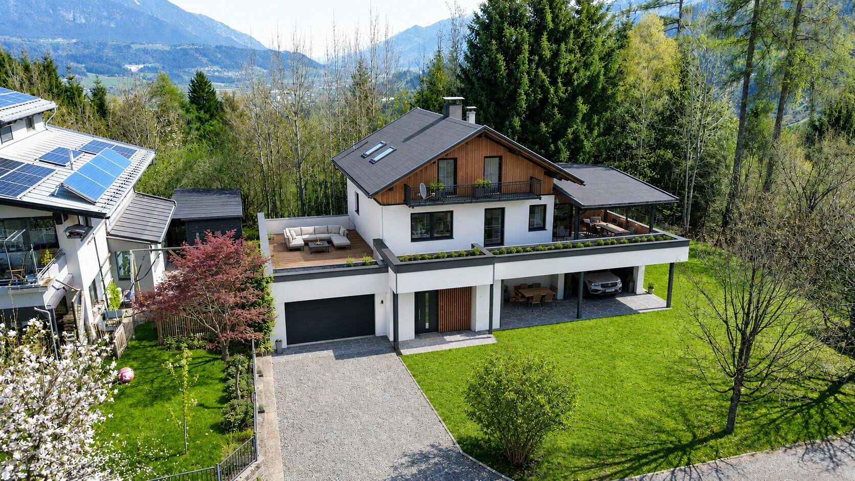 Modernes Wohnhaus mit weißer Fassade, Dachterrasse und Bergblick, umgeben von gepflegtem Garten.