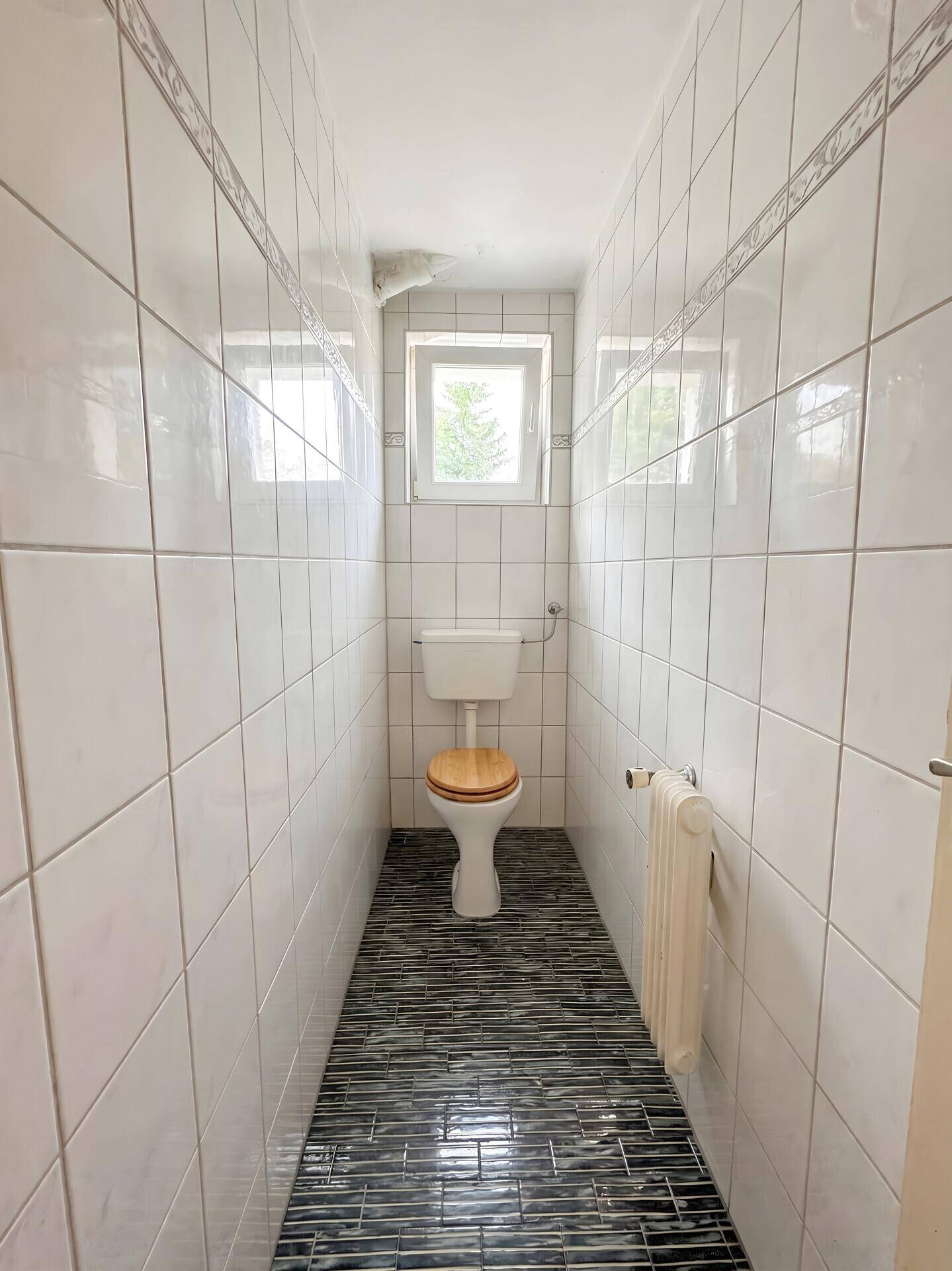 Separates WC mit Fenster, Toilette und Fliesenboden in schmalem Raum.