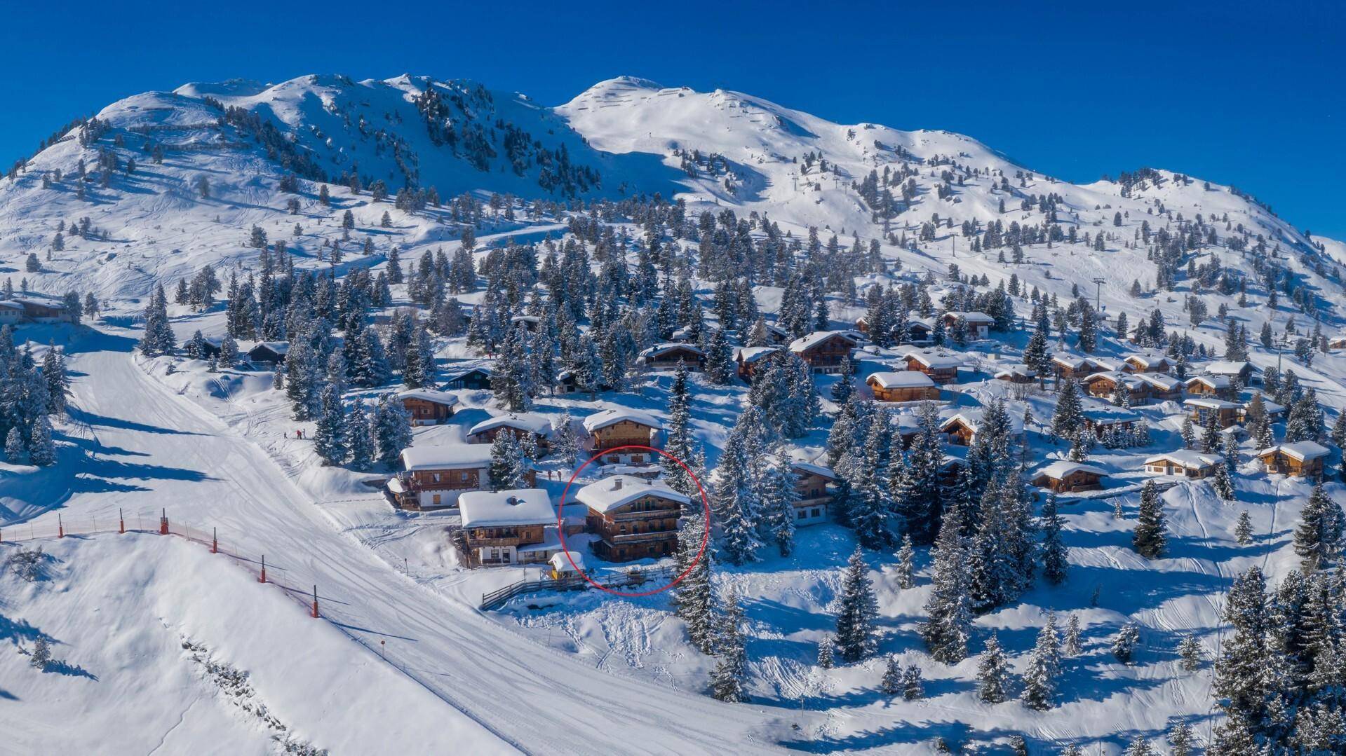 Luftaufnahme einer Ansammlung von Chalets in einer verschneiten Berglandschaft mit Skipiste.