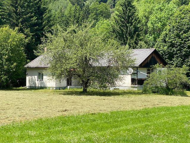 Nähere Ansicht des Hauses mit heller Fassade und dunklem Dach, umgeben von gemähten Wiesen und Bäumen.