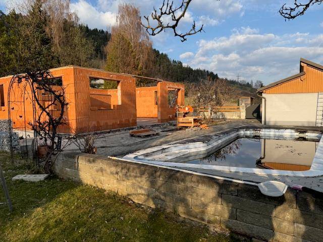 Rohbau eines Hauses mit Ziegelwänden und einem Poolbereich, umgeben von grüner Landschaft und Bäumen.