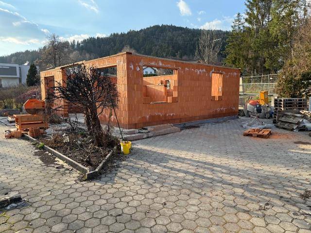 Seitenansicht des Rohbaus mit Ziegelwänden und großen Fensteröffnungen auf einem gepflasterten Grundstück.