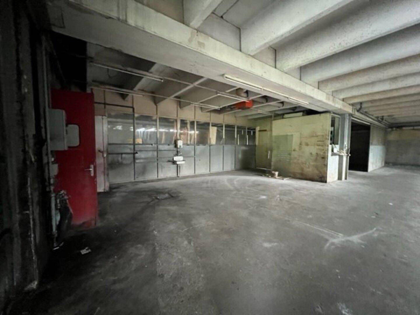Leere Industriehalle mit Betonboden, sichtbaren Deckenstrukturen und einer roten Tür an der Seite.
