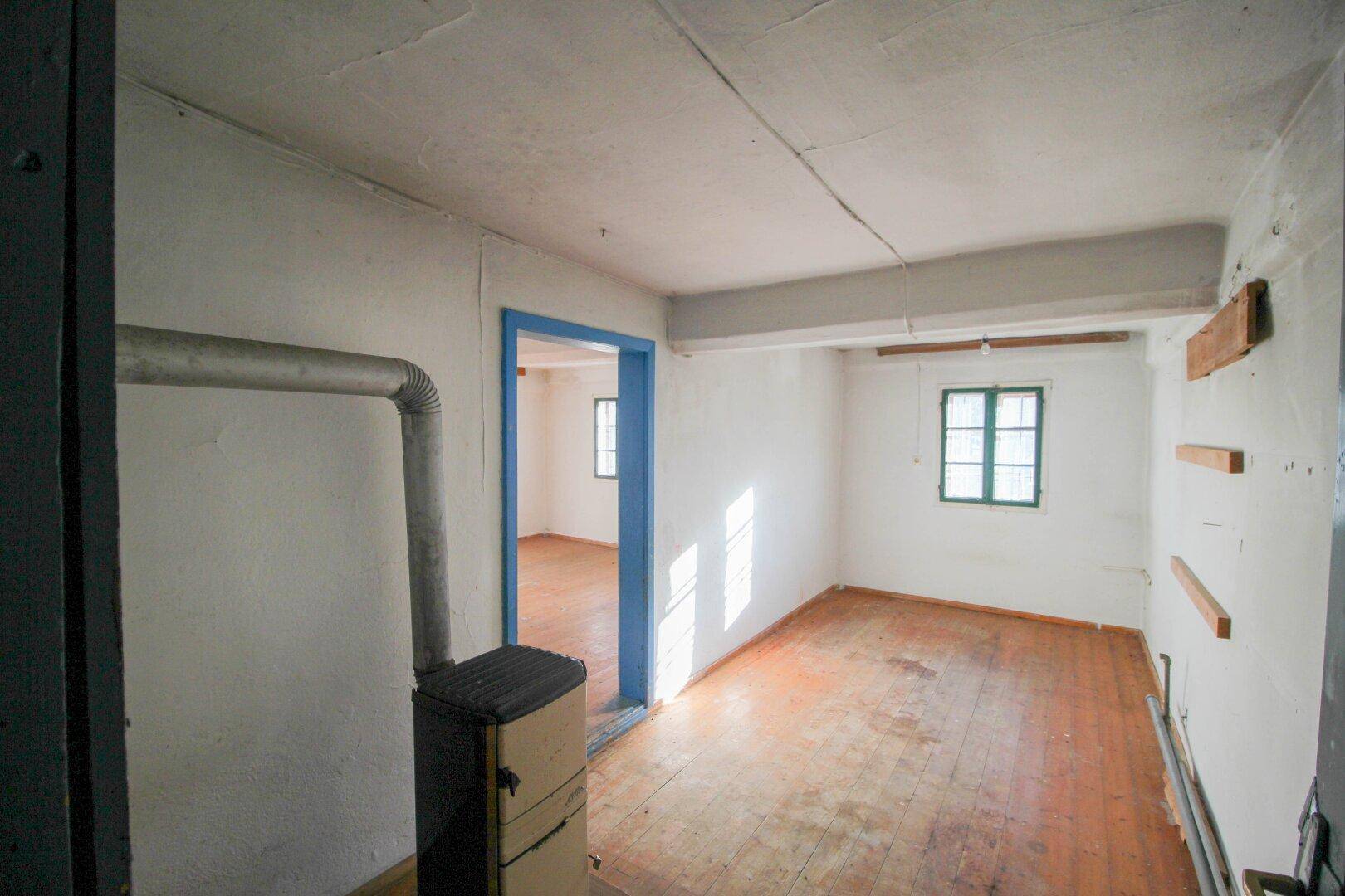 Leeres Zimmer mit Holzdielenboden, weiß gestrichenen Wänden und einem sichtbaren Holzofen. Ein Fenster mit grünen Rahmen lässt natürliches Licht herein.
