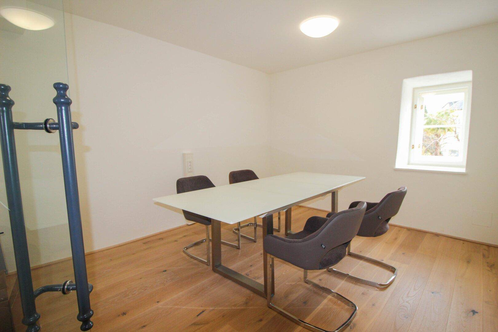 Heller Besprechungsraum mit Holzboden, weißem Tisch und modernen Stühlen, beleuchtet durch ein kleines Fenster.