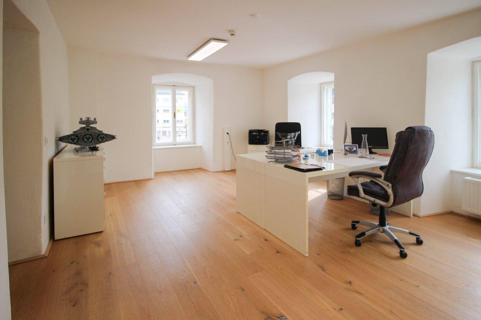 Großes Büro mit zwei Fenstern, hellem Holzboden, weißem Schreibtisch und Bürostühlen, sowie einem Drucker.
