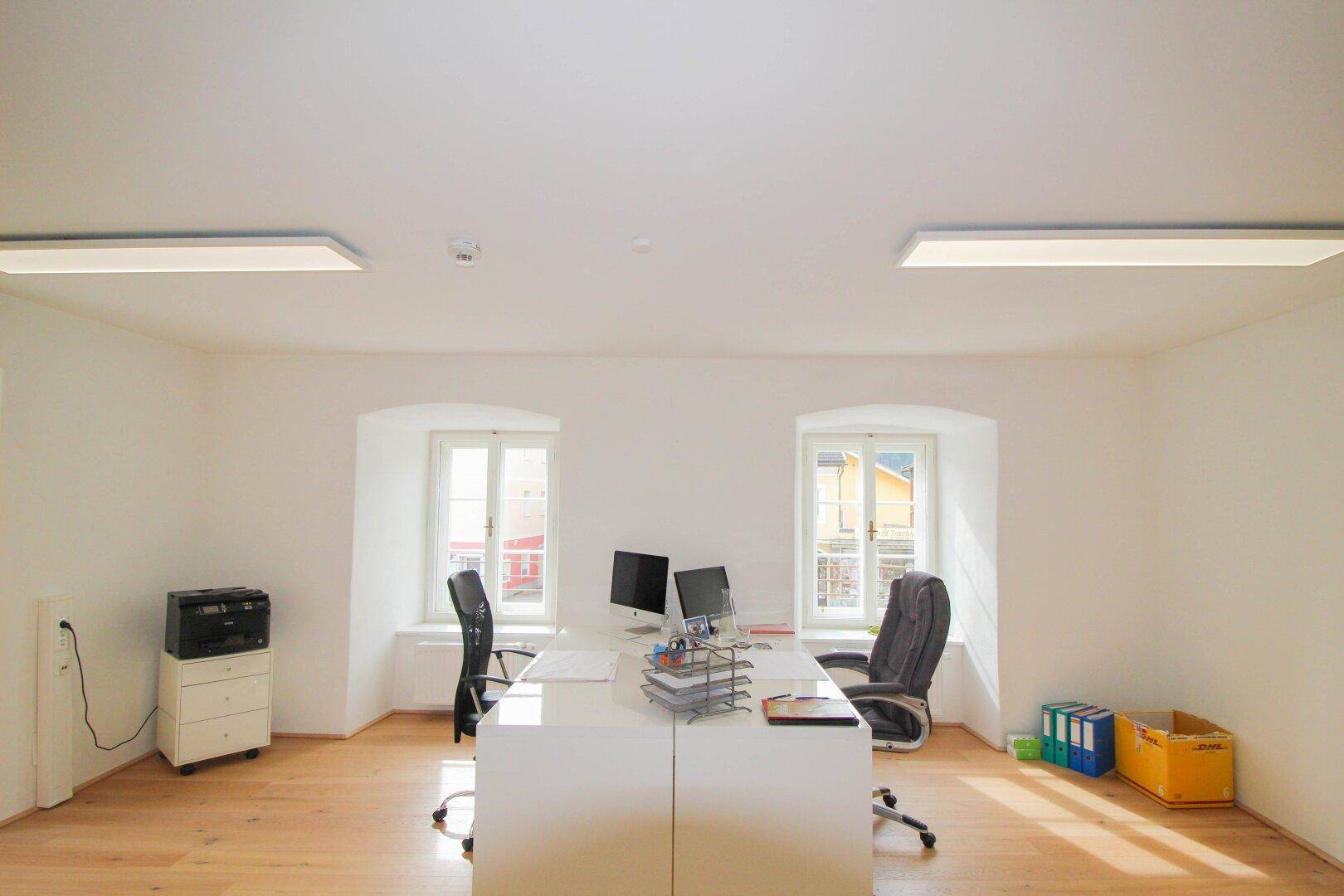 Helles Büro mit zwei Fenstern, großem Schreibtisch und Bürostühlen, sowie Deckenleuchten für zusätzliche Beleuchtung.