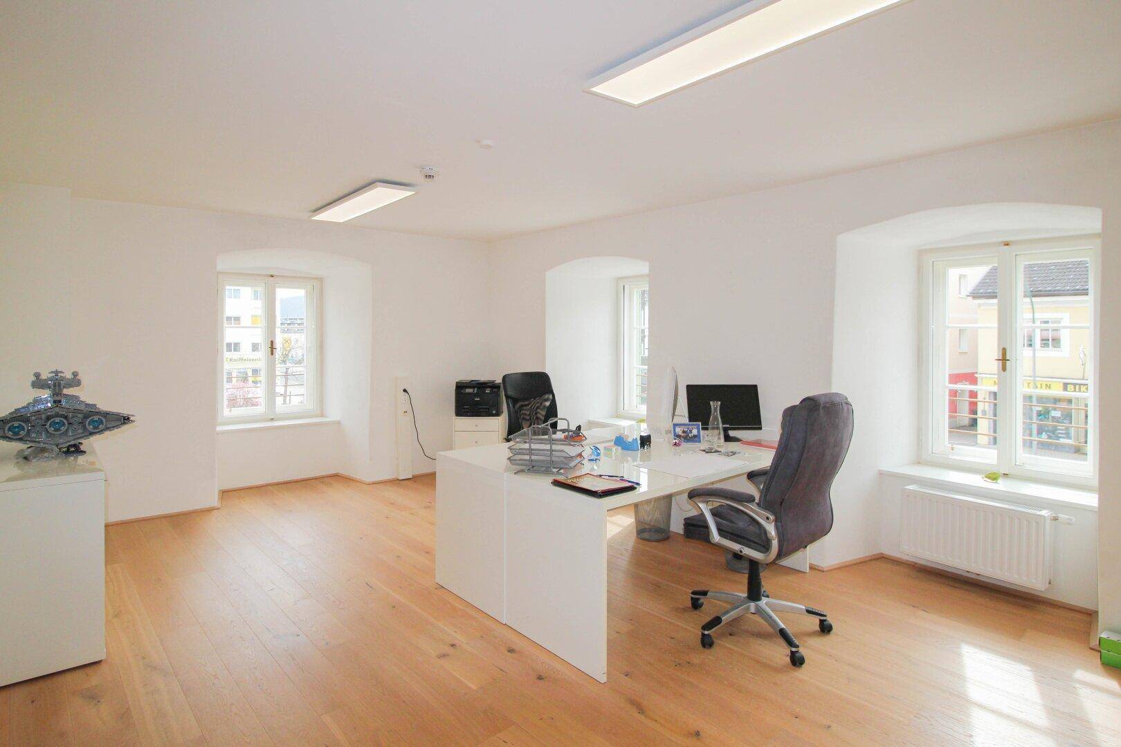 Geräumiges Büro mit hellem Holzboden, zwei Fenstern und einem großen weißen Schreibtisch mit Bürostühlen.