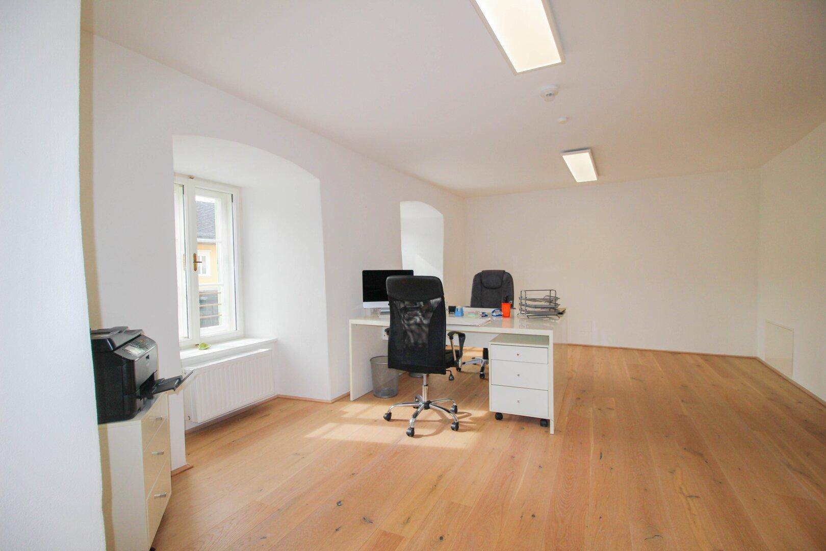 Geräumiges Büro mit hellem Holzboden, weißem Schreibtisch und Drucker, gut beleuchtet durch ein Fenster.