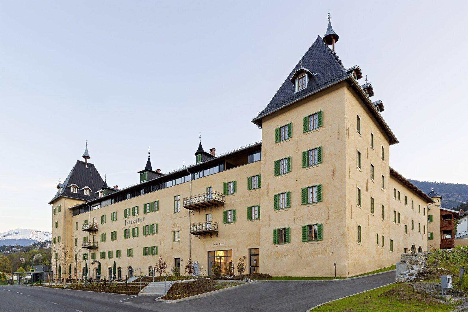Historisches Gebäude mit markanten Türmen und grünen Fensterläden, umgeben von einer gepflegten Außenanlage.