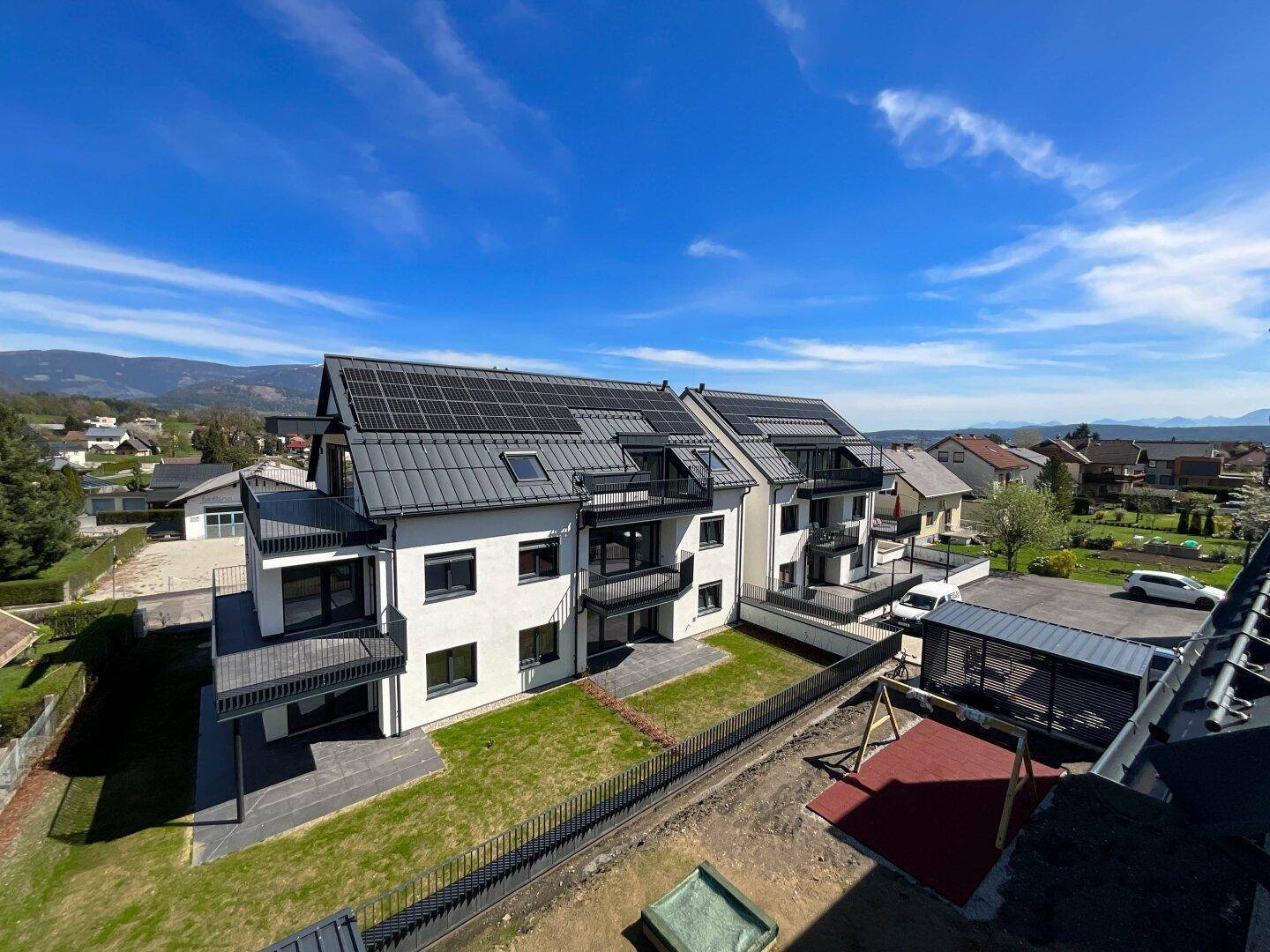 Moderne Wohnanlage mit weißen Fassaden, schwarzen Dächern, Solarpaneelen und Bergblick.