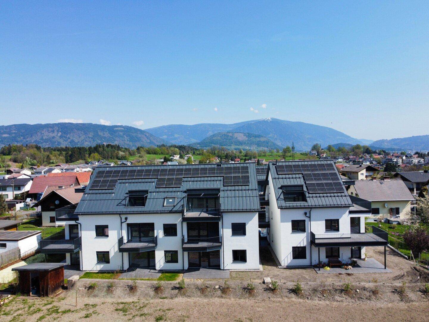 Zwei moderne Wohngebäude mit Photovoltaikanlagen auf den Dächern und Bergblick in sonniger Umgebung.