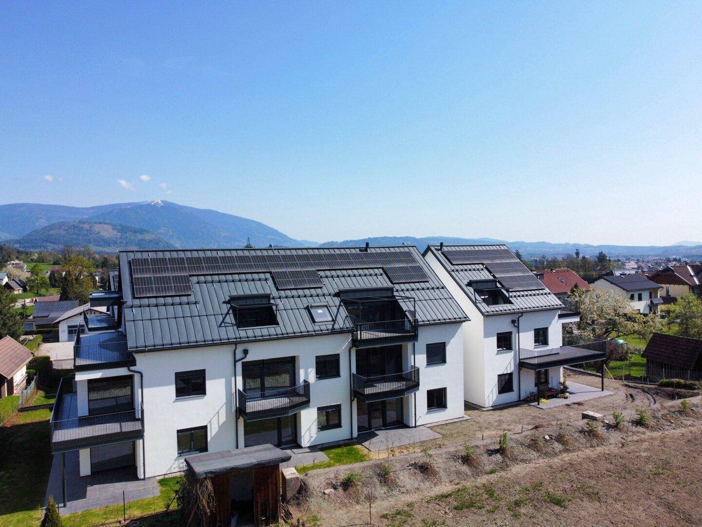 Neue Wohngebäude mit weißer Fassade, dunklen Dächern und integrierten Solarmodulen vor Bergkulisse.