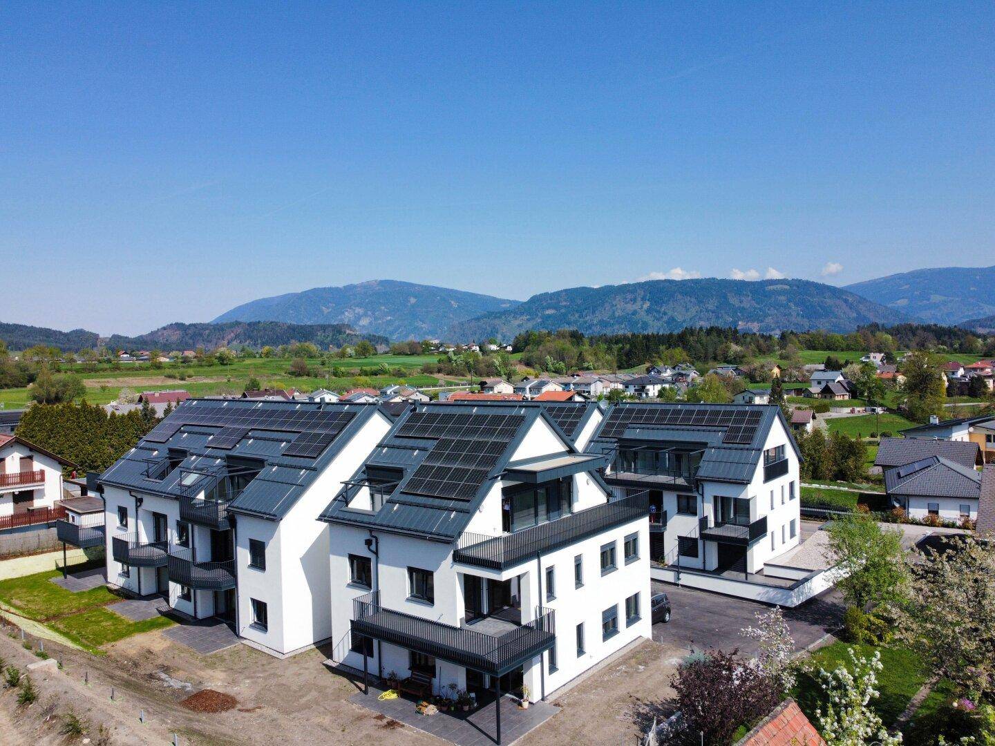 Drei moderne Wohngebäude mit klaren Linien, Photovoltaikanlagen und Bergblick in sonniger Lage.