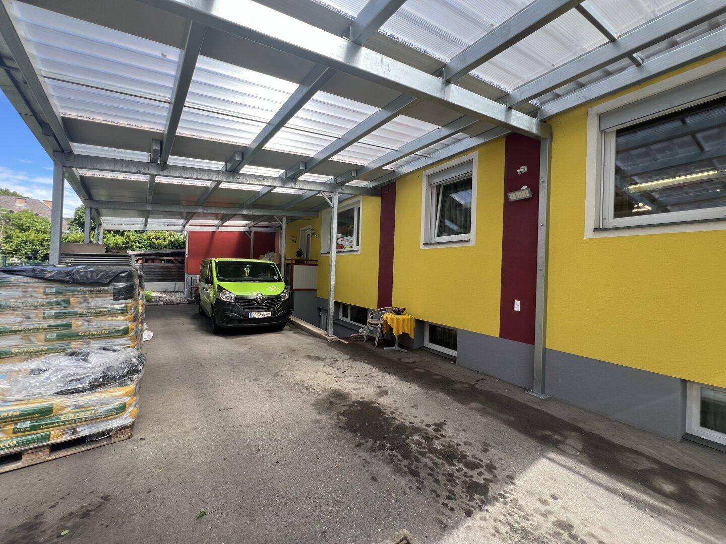 Überdachter Bereich neben einem gelben Gebäude mit einem Lieferwagen und gestapelten Paletten.