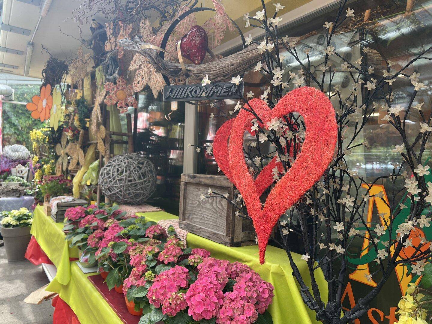 Nahaufnahme einer bunten Blumenauslage mit roten Herzen und einem "Willkommen"-Schild im Schaufenster.