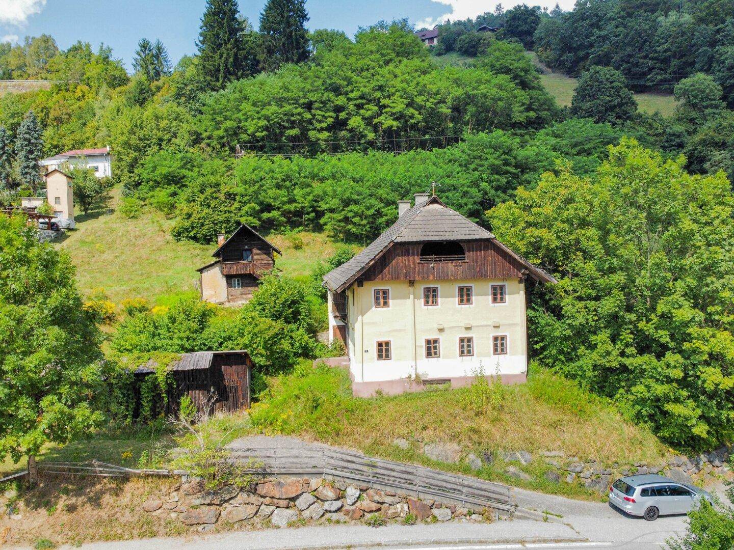 Traditionelles Haus in alpiner Landschaft mit Holzbalkon und umliegenden Wäldern, inklusive Parkplatz.