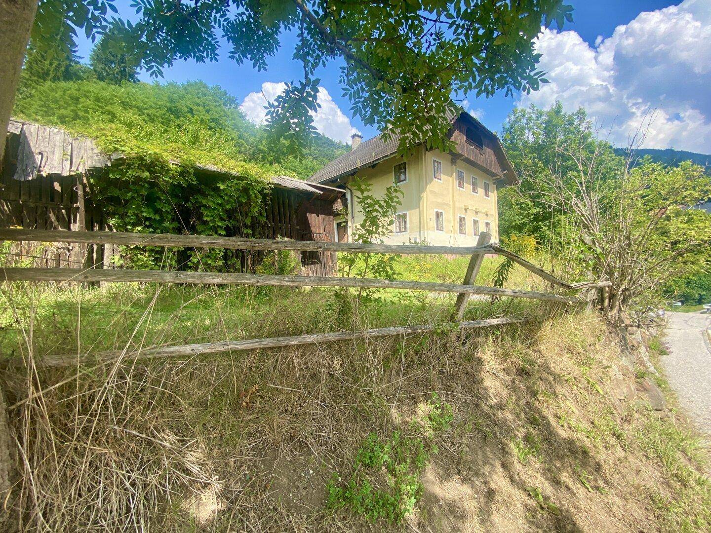 Seitenansicht des Hauses, umgeben von üppigem Grün und einem rustikalen Holzzaun auf einem Hanggrundstück.