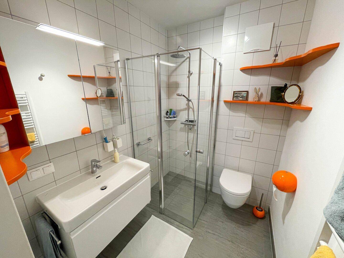 Modernes Badezimmer mit weißen Fliesen, begehbarer Dusche, wandhängendem WC und farbigen Akzenten.