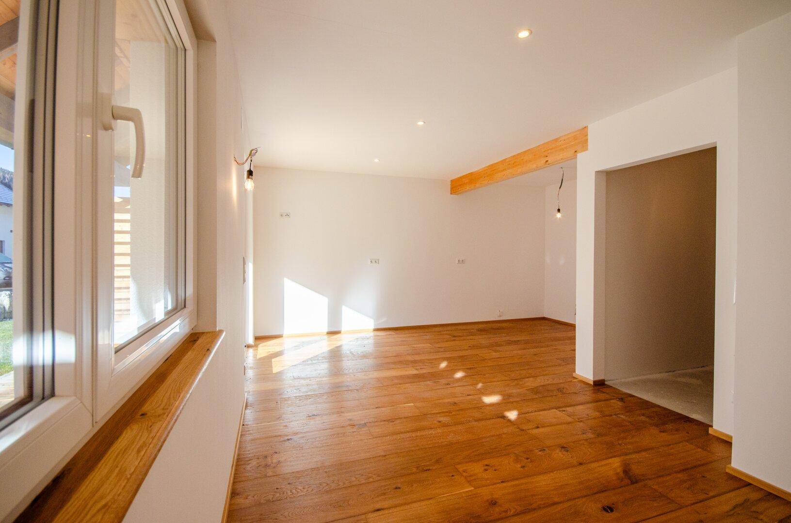 Heller, leerer Raum mit Holzboden und einem großen Fenster, das viel Tageslicht hereinlässt.