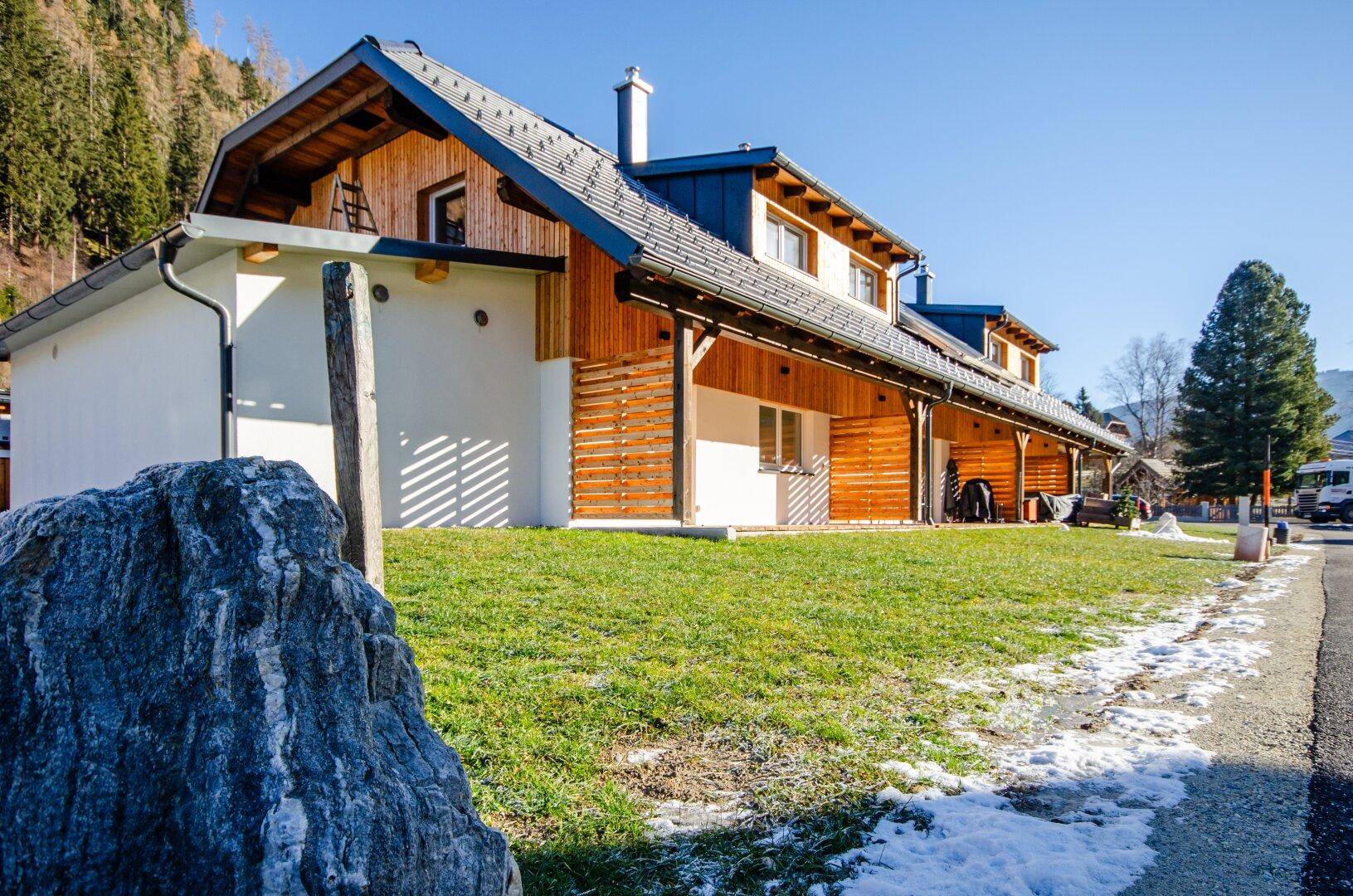 Modernes Gebäude mit Holzfassade und weißem Putz, umgeben von Rasen und Bergen im Hintergrund.